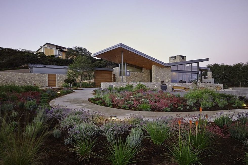 Modern house with sloped roof, stone walls, and lush garden at dusk.