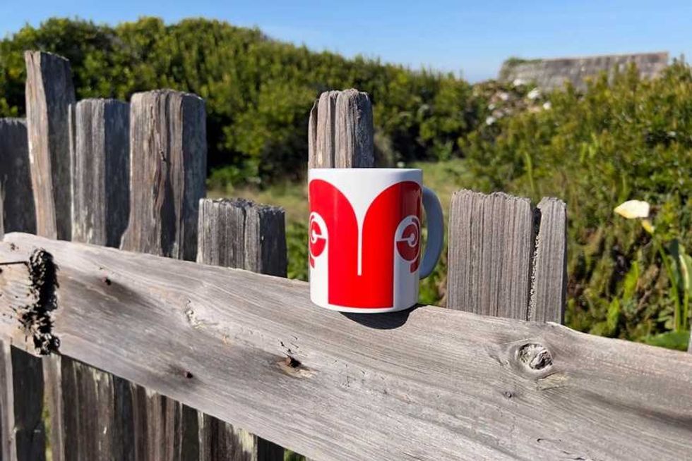 Mug with red design on rustic wooden fence, green bushes in background.