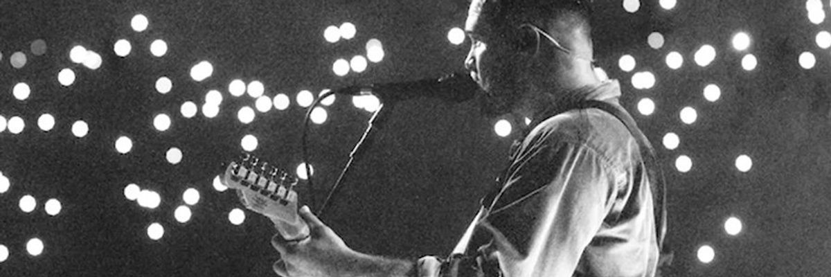 Musician plays guitar on stage, with microphone and bright lights in the background.