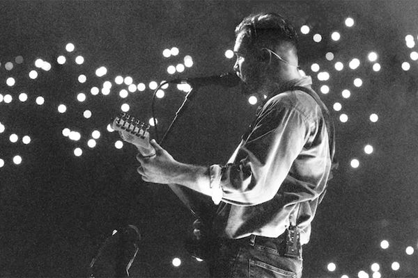 Musician plays guitar on stage, with microphone and bright lights in the background.