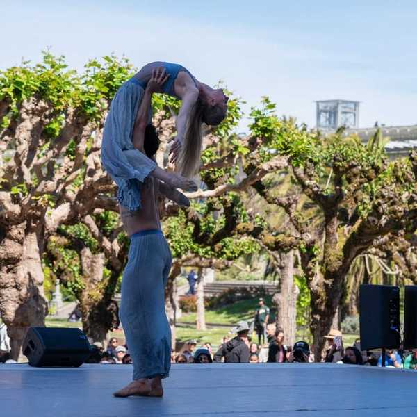 One dancer lifting another on stage in a park