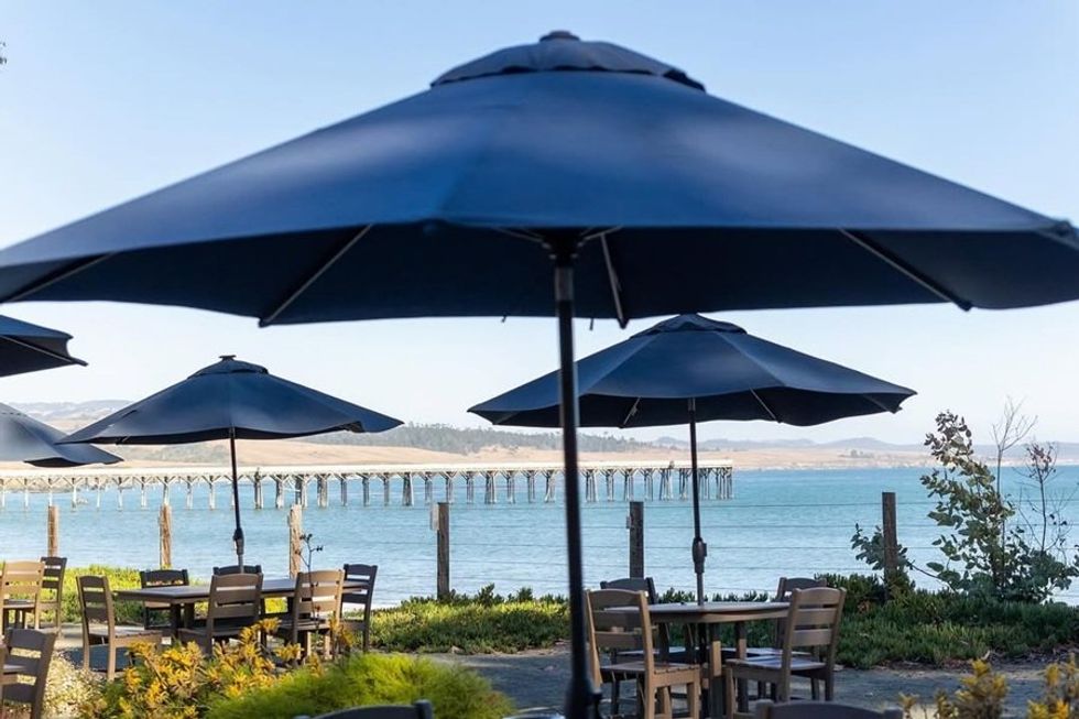 Outdoor cafe with blue umbrellas by the sea, overlooking a pier and distant hills.