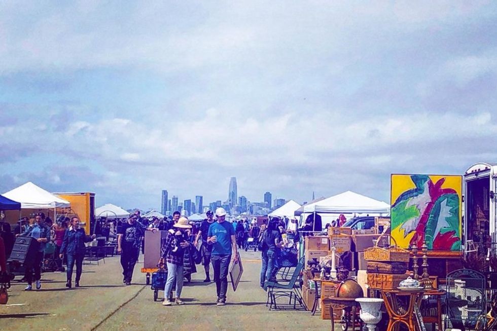 Outdoor market with people browsing, city skyline in background.