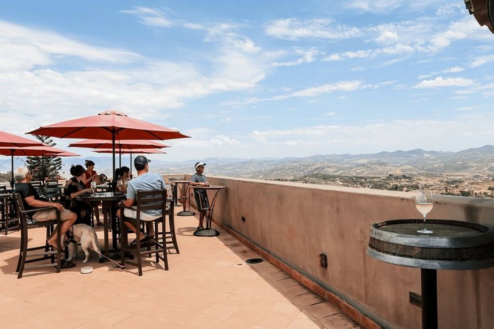Outdoor patio with red umbrellas, people dining, overlooking scenic mountains.