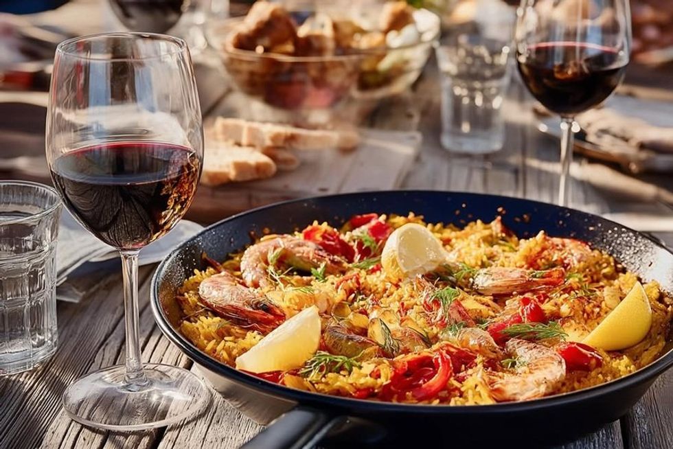 Paella with seafood and lemon wedges beside a glass of red wine on a rustic table.