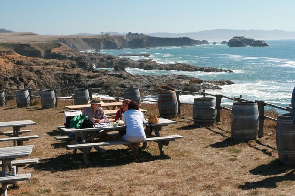 People at a picnic table by the ocean, surrounded by barrels and rugged cliffs.