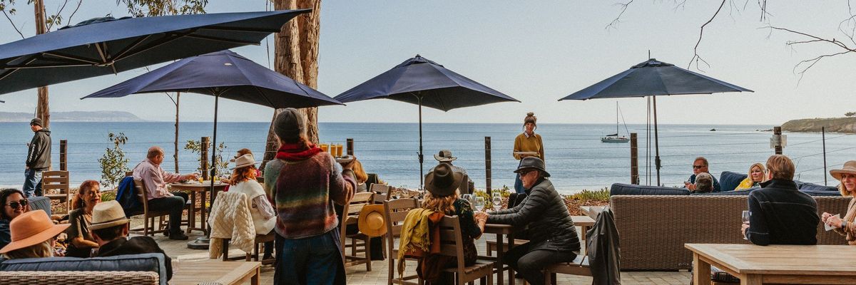 People dining outdoors by the sea under blue umbrellas and trees.