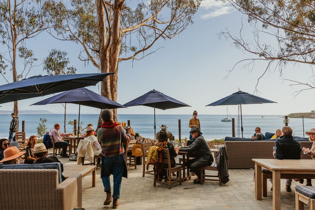 People dining outdoors by the sea under blue umbrellas and trees.