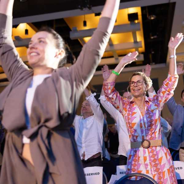 people in a conference audience on their feet with their hands raised and smiling