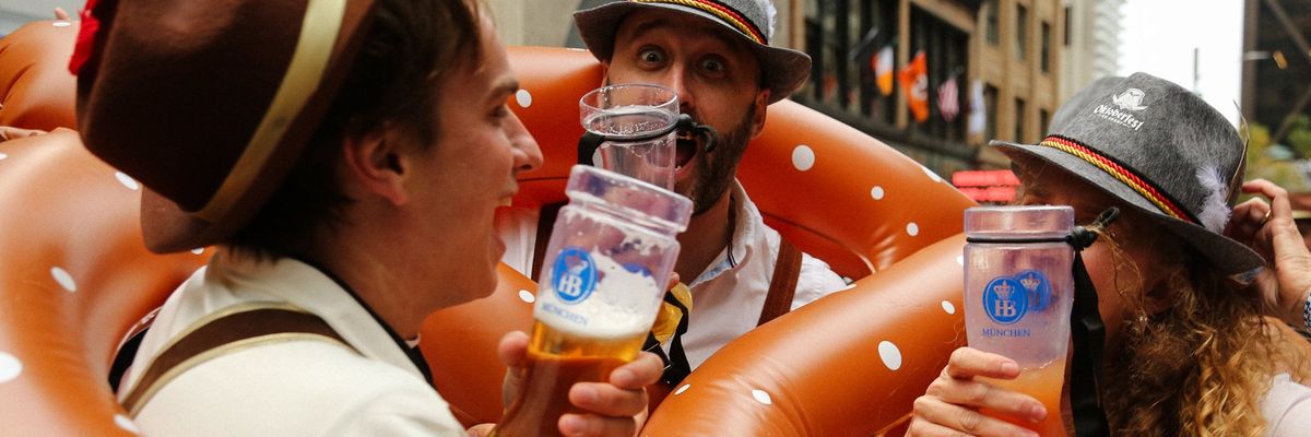 People in Bavarian hats drink beer, surrounded by inflatable pretzels at a festive event.