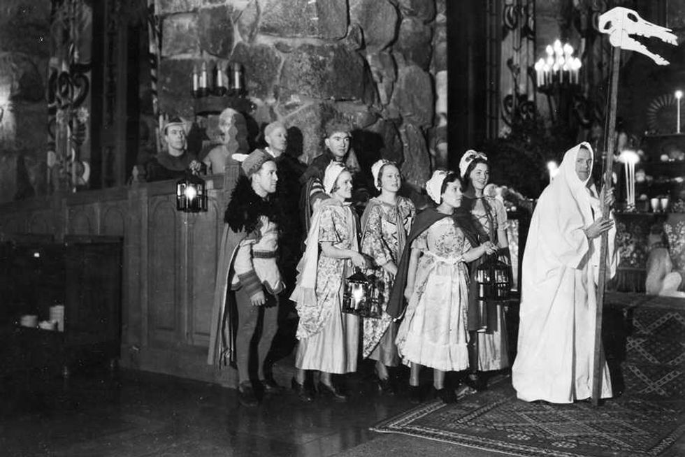 People in historical costumes with lanterns gather indoors near a robed figure holding a staff.
