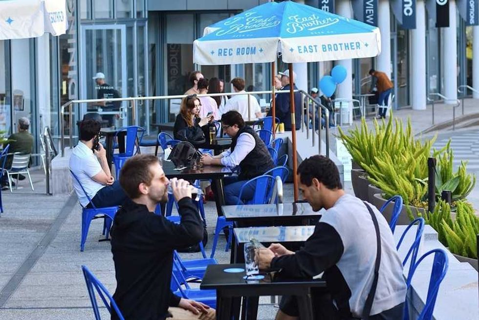 People sitting at outdoor tables under umbrellas at a cafe, with blue chairs and plants.