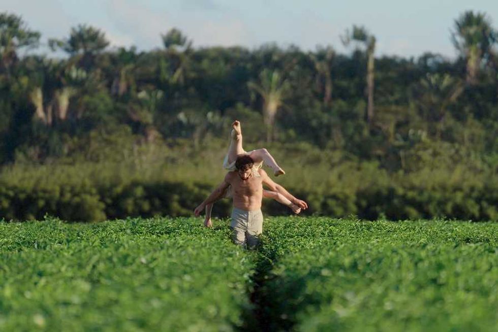 Person carrying another through a green field, surrounded by tall trees.