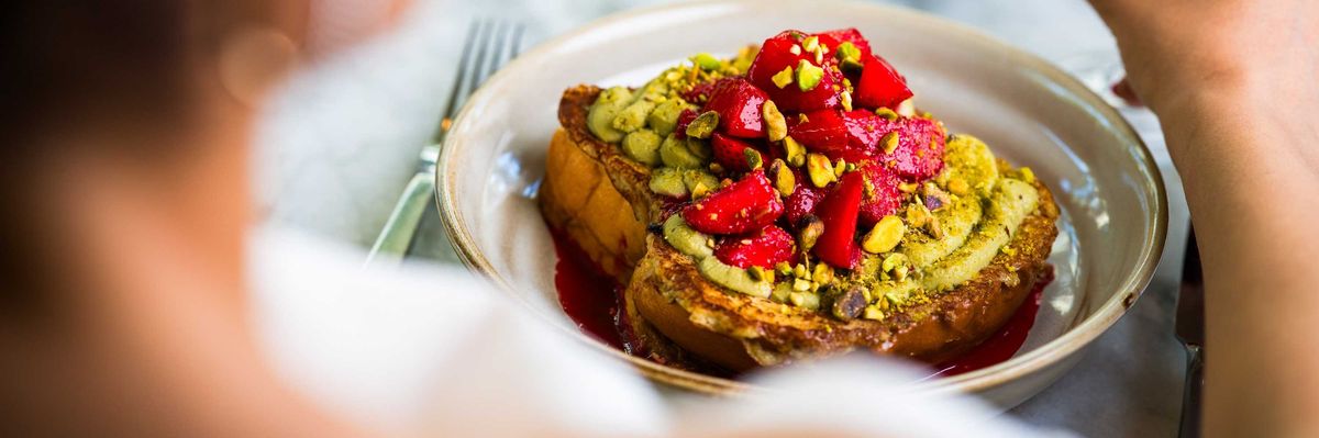 Person eating toast topped with strawberries and pistachios, holding a berry smoothie.
