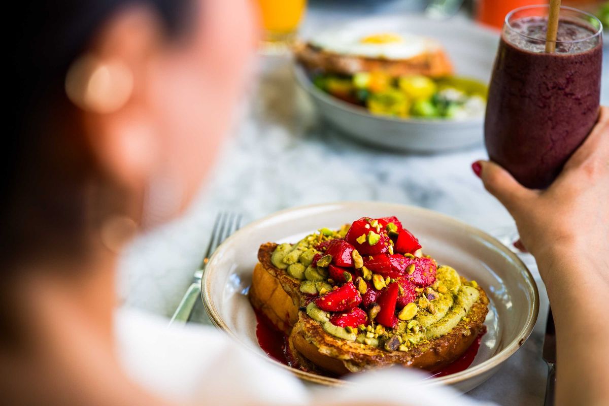 Person eating toast topped with strawberries and pistachios, holding a berry smoothie.