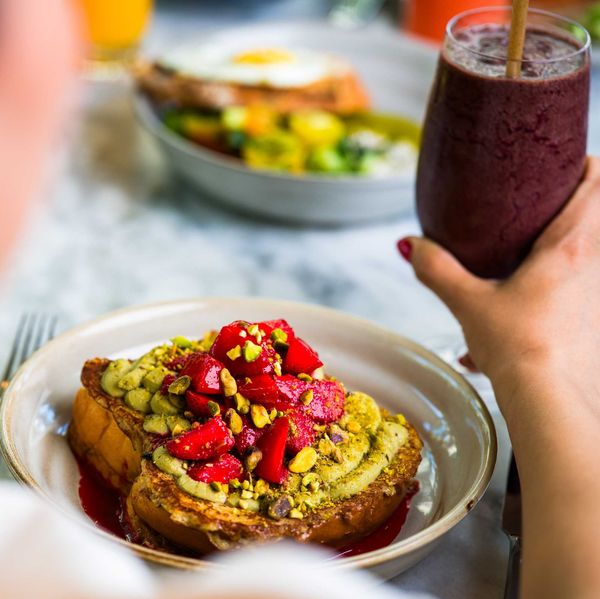 Person eating toast topped with strawberries and pistachios, holding a berry smoothie.