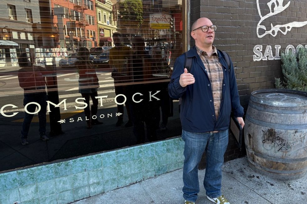 Person gesturing outside Comstock Saloon with barrel and building reflections nearby.