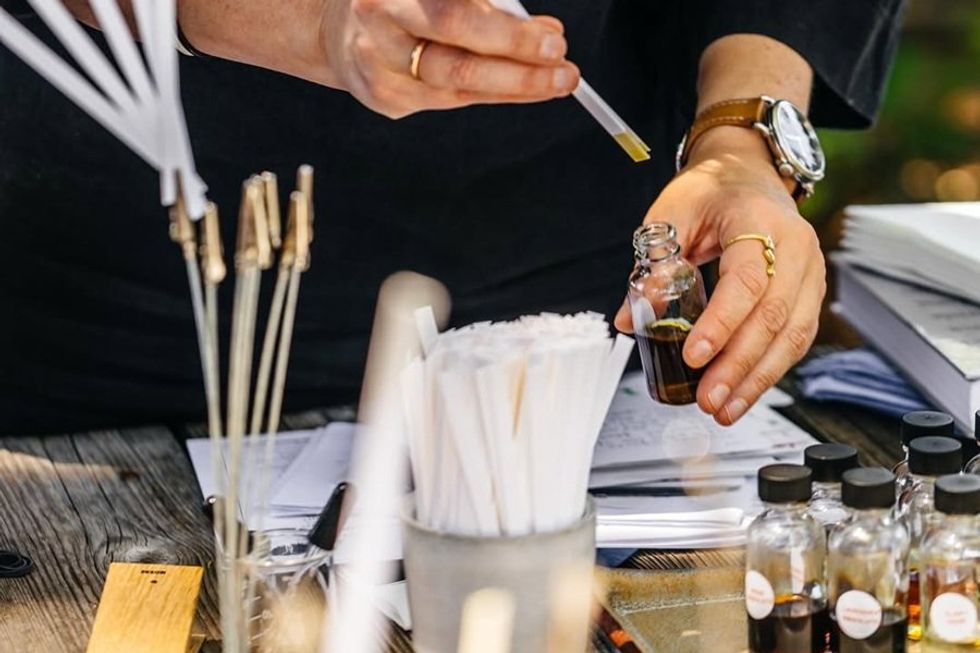 Person handling a pipette and bottle near scent testing strips on a wooden table.