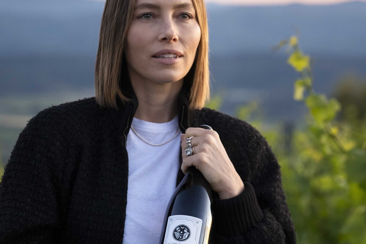 Person holding a wine bottle outdoors, vineyard in the background.