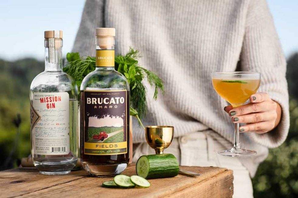 Person holding cocktail near gin bottles, herbs, and sliced cucumber on a wooden table.