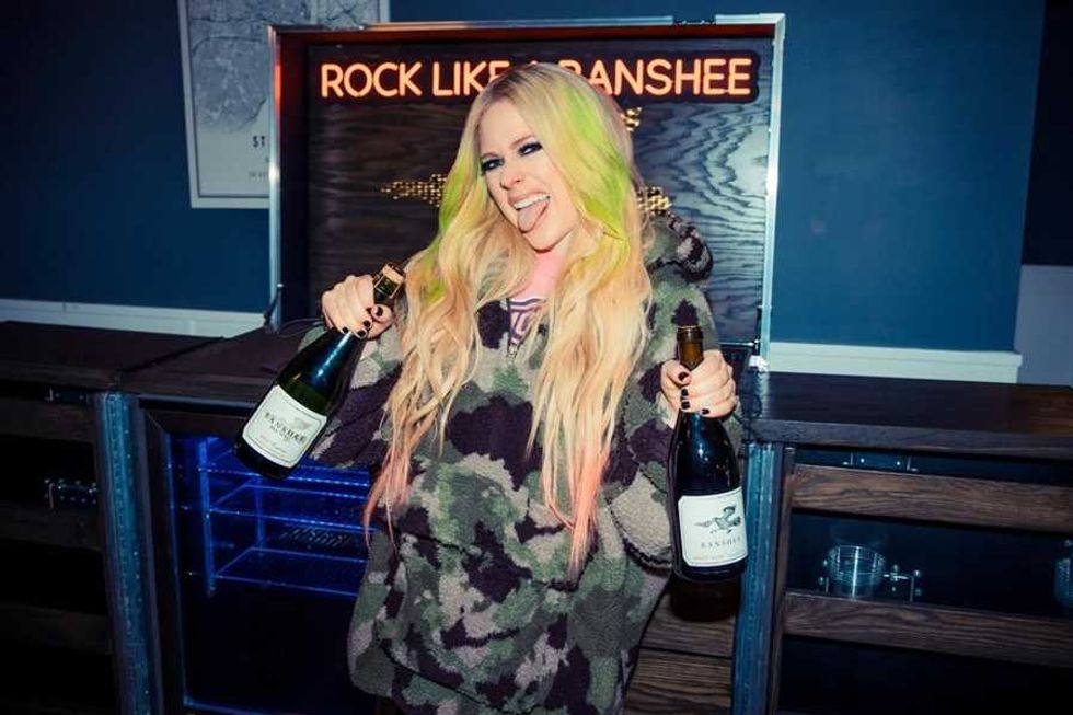 Person in a camo hoodie holding two wine bottles, tongue out, with "Rock Like a Banshee" sign.