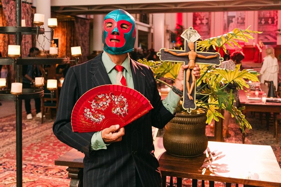 Person in a luchador mask holding a red fan and crucifix in a colorful, candle-lit room.