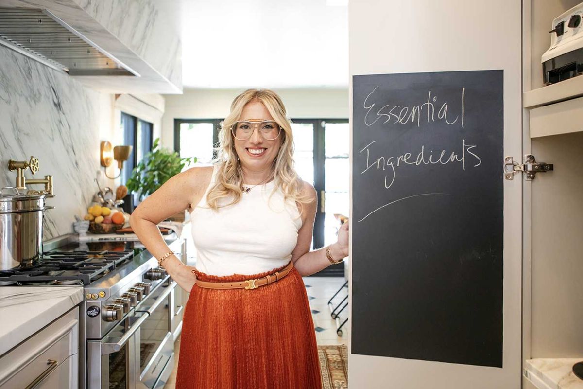 Person in kitchen by chalkboard labeled "Essential Ingredients," smiling in a white top and red skirt.