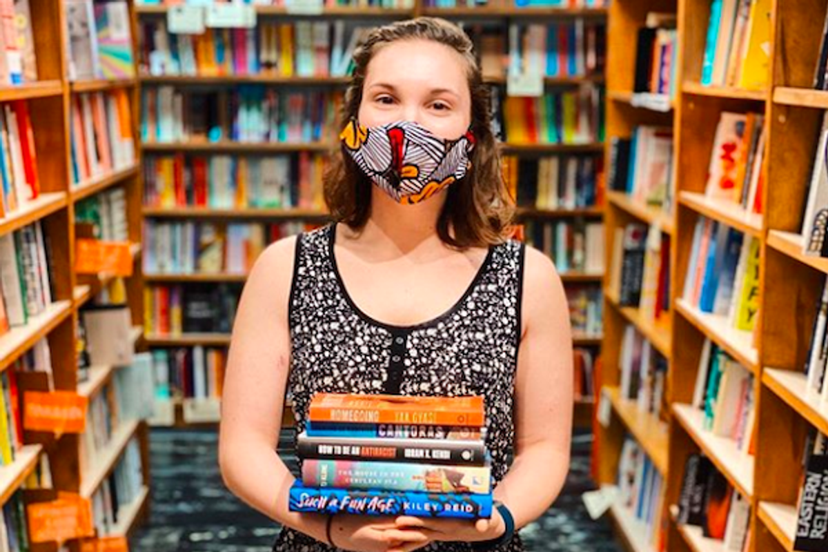 Person in mask holding books in a colorful bookstore aisle.