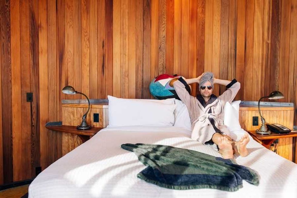 Person in robe and sunglasses relaxing on bed, wood-paneled room, towel on the bed.