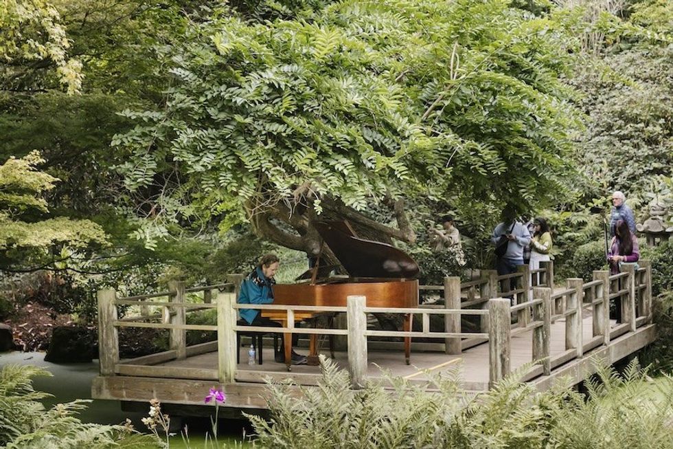 Person plays piano on a wooden platform surrounded by lush greenery and people.