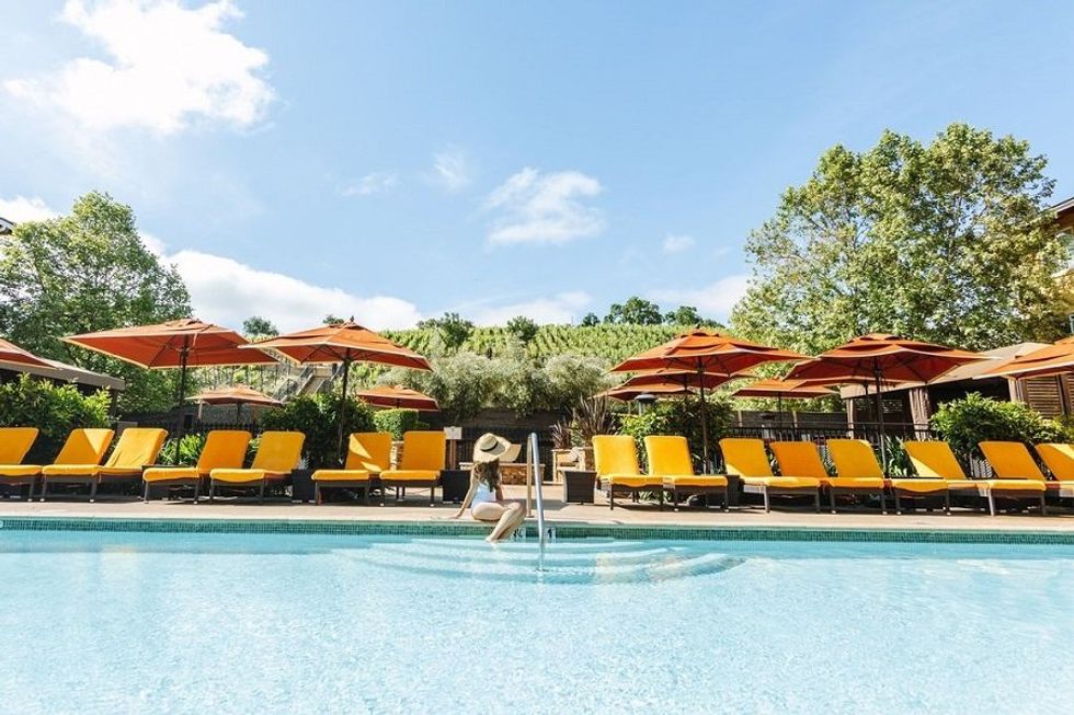 Person relaxing by a pool, surrounded by orange umbrellas and loungers on a sunny day.