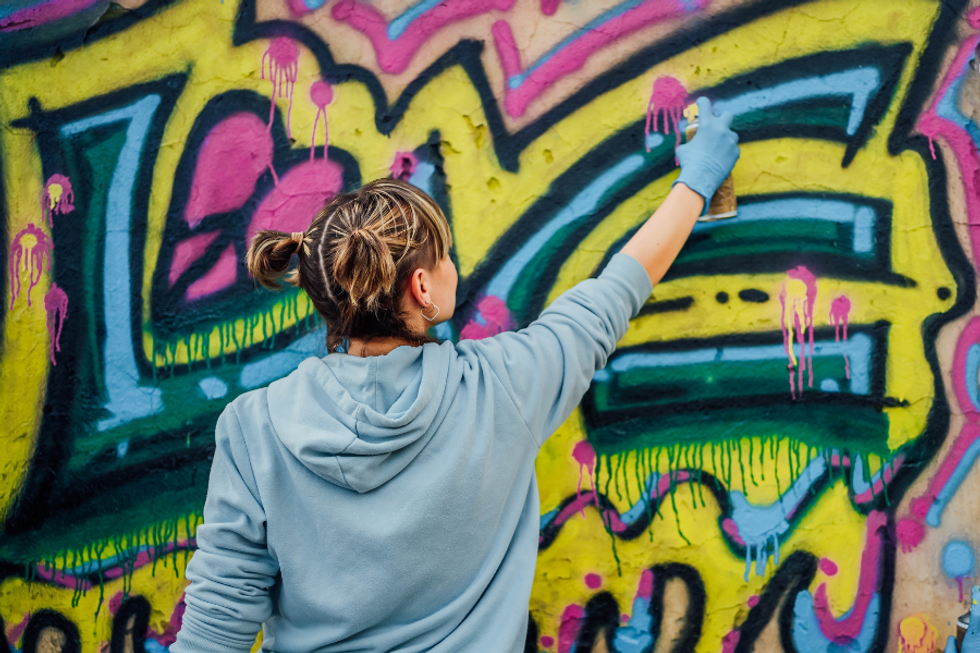 Person spray painting colorful graffiti on a wall, wearing a hoodie and blue glove.
