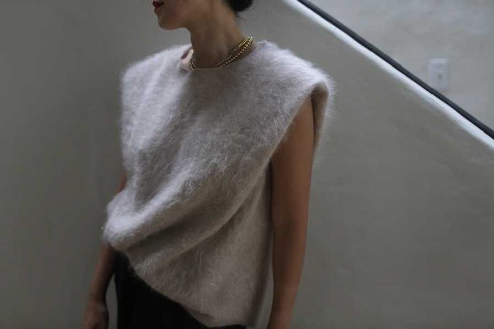 Person wearing a fuzzy sleeveless top and gold necklace, standing by a white wall.