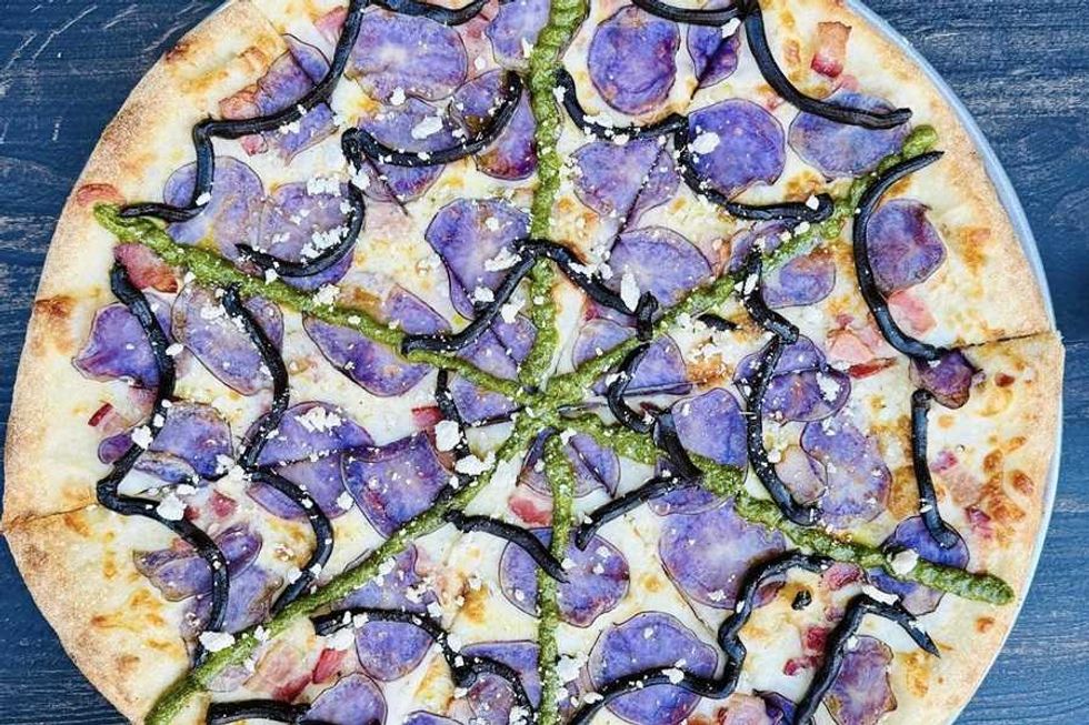 Pizza topped with purple potatoes, black and green drizzles, and cheese on a wooden table.
