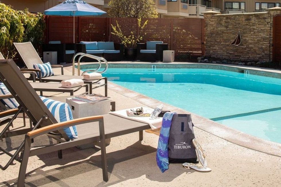 Poolside with lounge chairs, towels, a tote bag, and blue parasol under sunny skies.