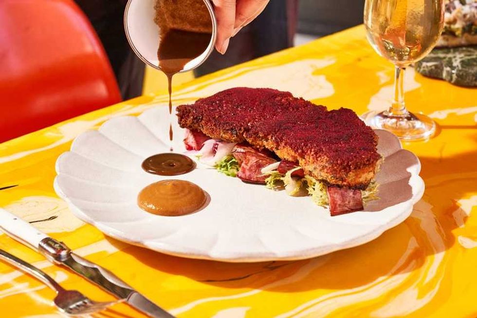 Pouring sauce over crispy coated steak on a white plate with greens.