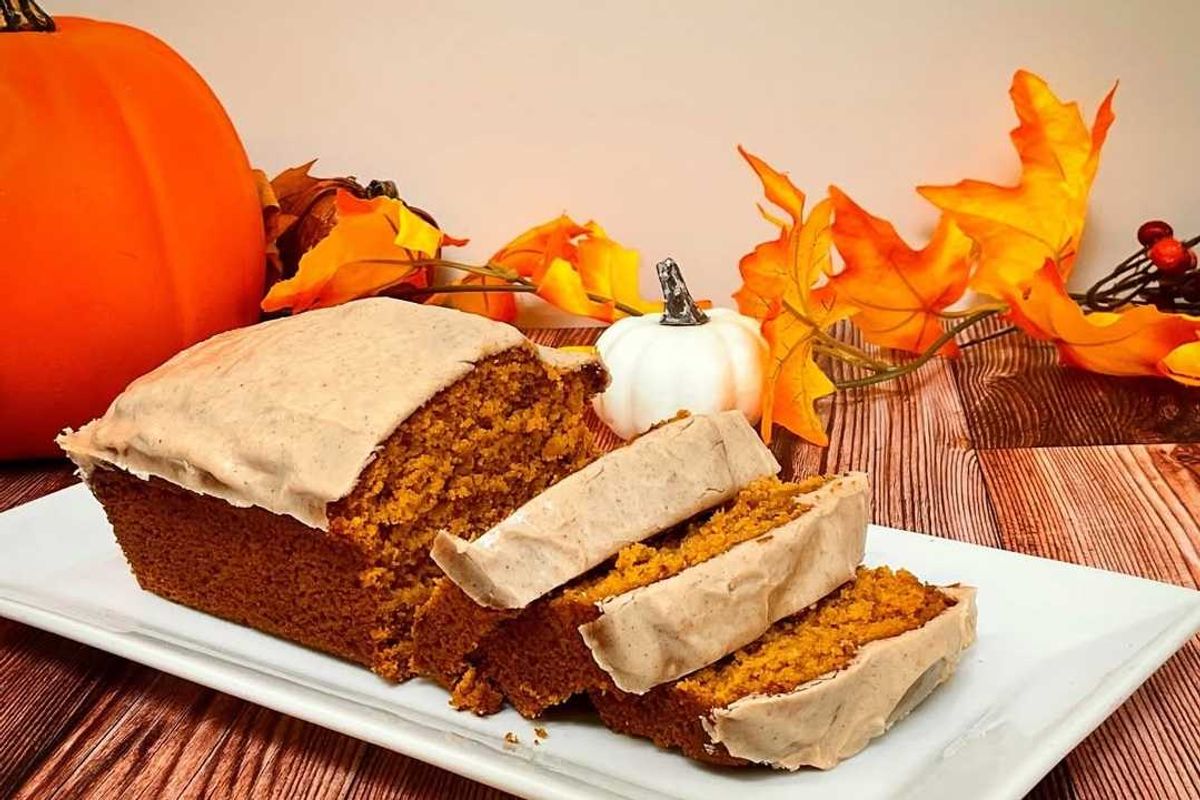 Pumpkin bread with frosting, surrounded by pumpkins and autumn leaves on a wooden table.
