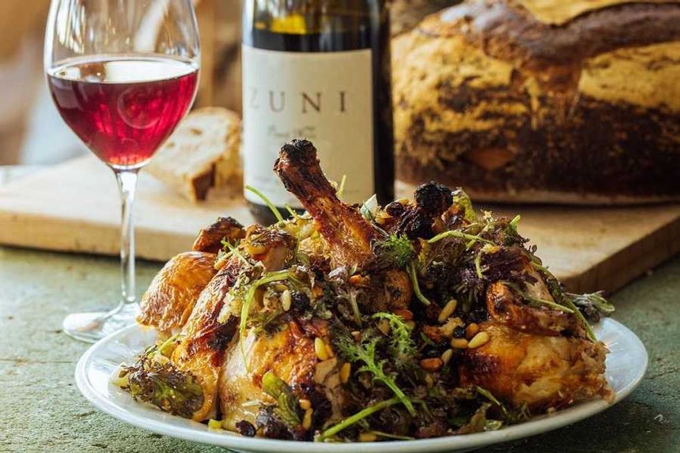 Roasted chicken with herbs, glass of red wine, and loaf of bread on a table.