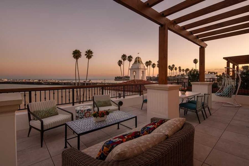 Rooftop patio with seating, pergola, and ocean view at sunset, surrounded by palm trees.