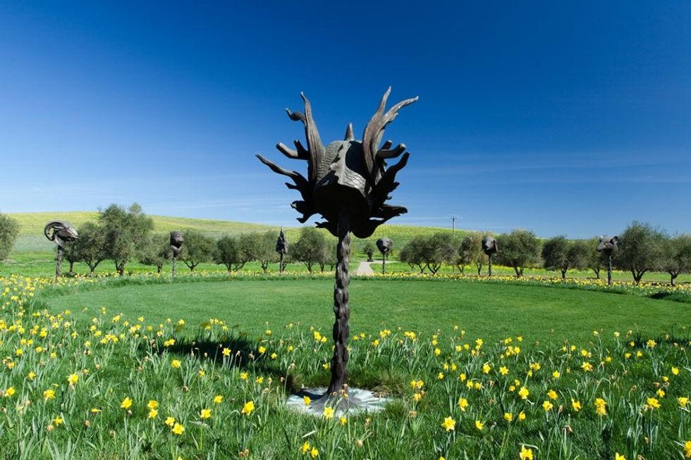 Sculptures circle in grassy field with yellow flowers and trees, under clear blue sky.