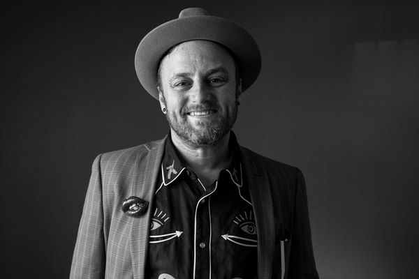 Smiling man in a hat and patterned shirt, wearing a plaid jacket, in black and white.
