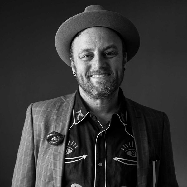 Smiling man in a hat and patterned shirt, wearing a plaid jacket, in black and white.