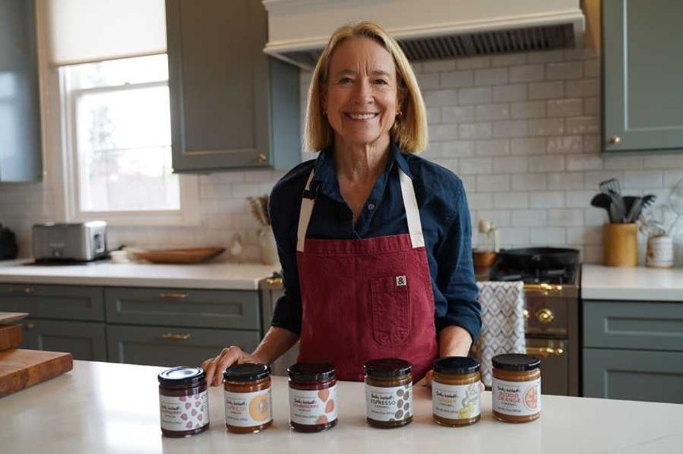 Smiling person in kitchen with six jars of spreads on the counter.