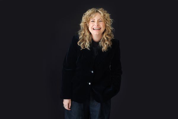 Smiling person with curly hair in a black jacket against a dark background.