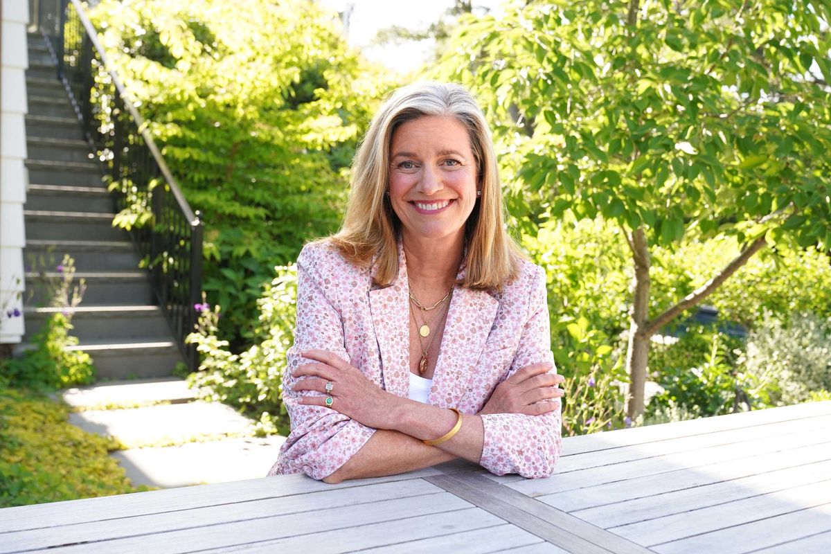 Smiling woman in a garden sitting at a table, wearing a floral-patterned blazer.