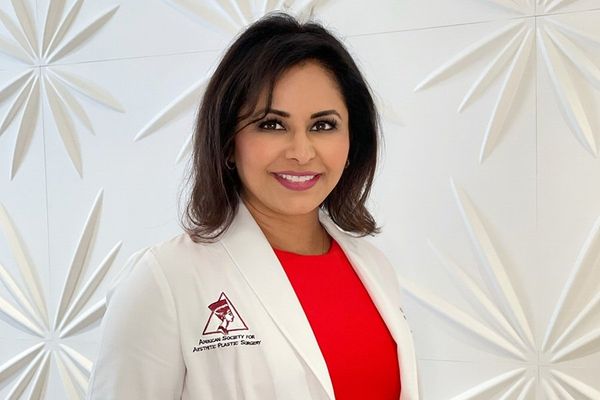 Smiling woman in a white coat stands against a textured white wall.