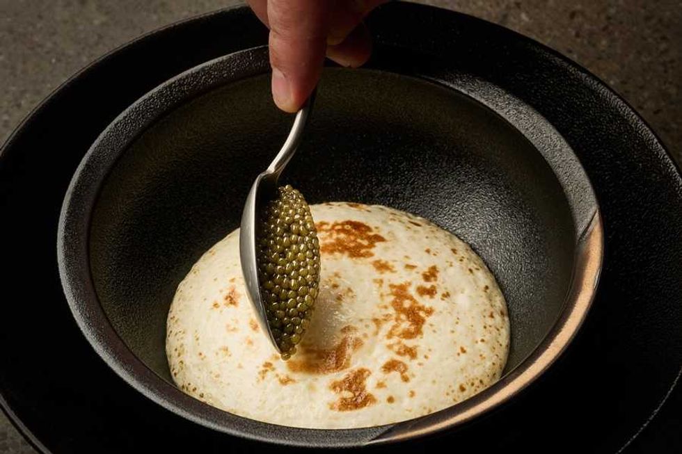 Spoon with caviar over a fluffy pancake in a black bowl.