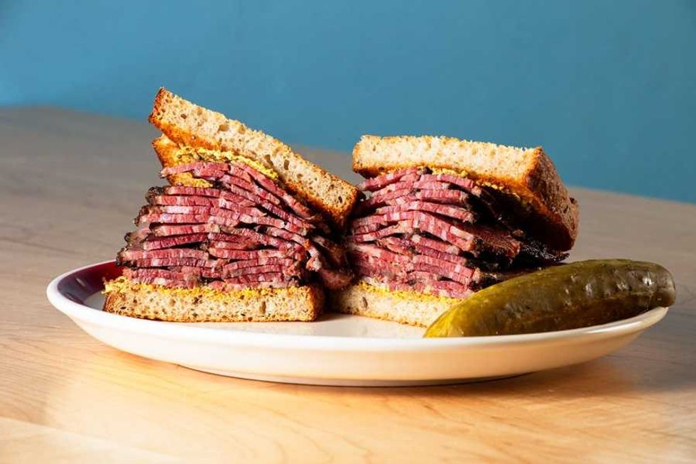 Stacked pastrami sandwich on rye with mustard, served with a pickle on a white plate.
