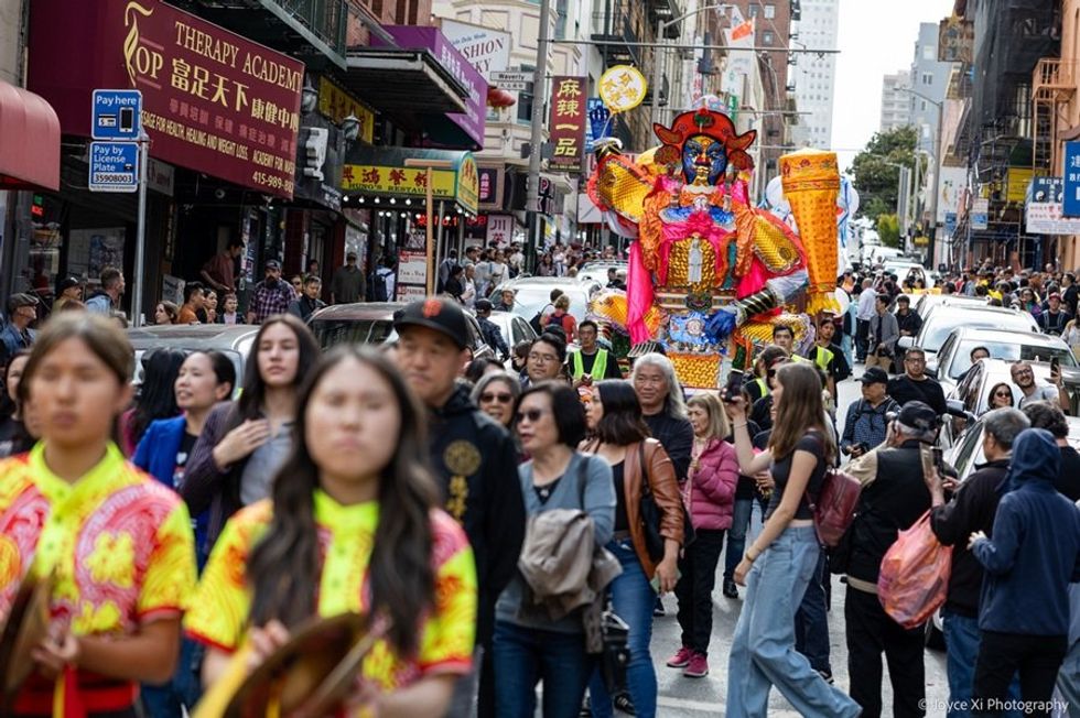 Street parade with a colorful deity float, crowded with spectators and performers.