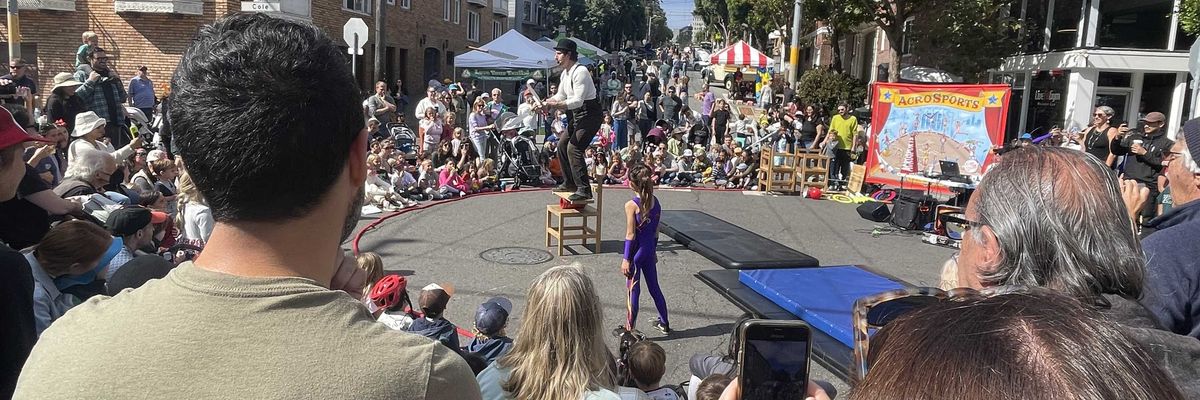 Street performance with a crowd watching a balancing act in a sunny urban setting.